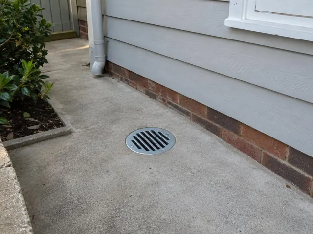 Outdoor grated drain set in dry concrete paving beside a house wall in Rose Bay